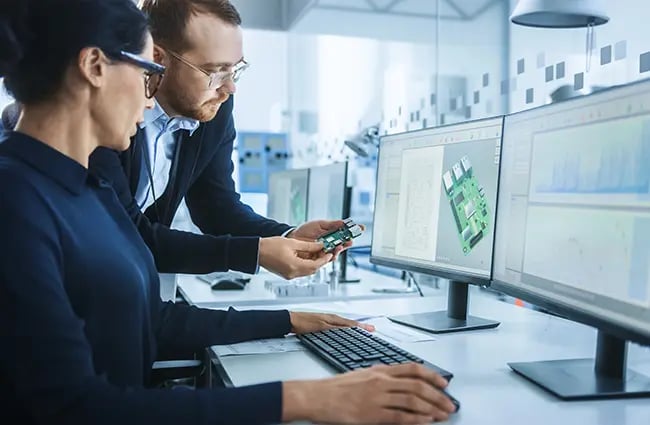 Two engineers reviewing a device in front of computer monitors