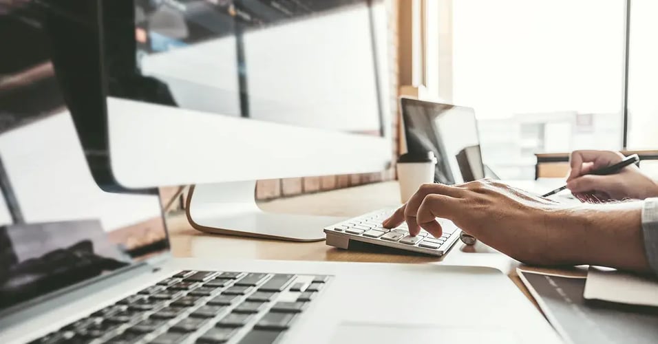 Hands typing at a keyboard in front of a computer and laptop.